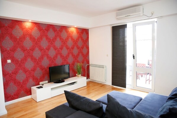 A modern living room with a red patterned wall, a flat-screen TV on a white entertainment unit, a radiator, and a sliding glass door leading to an outdoor area.