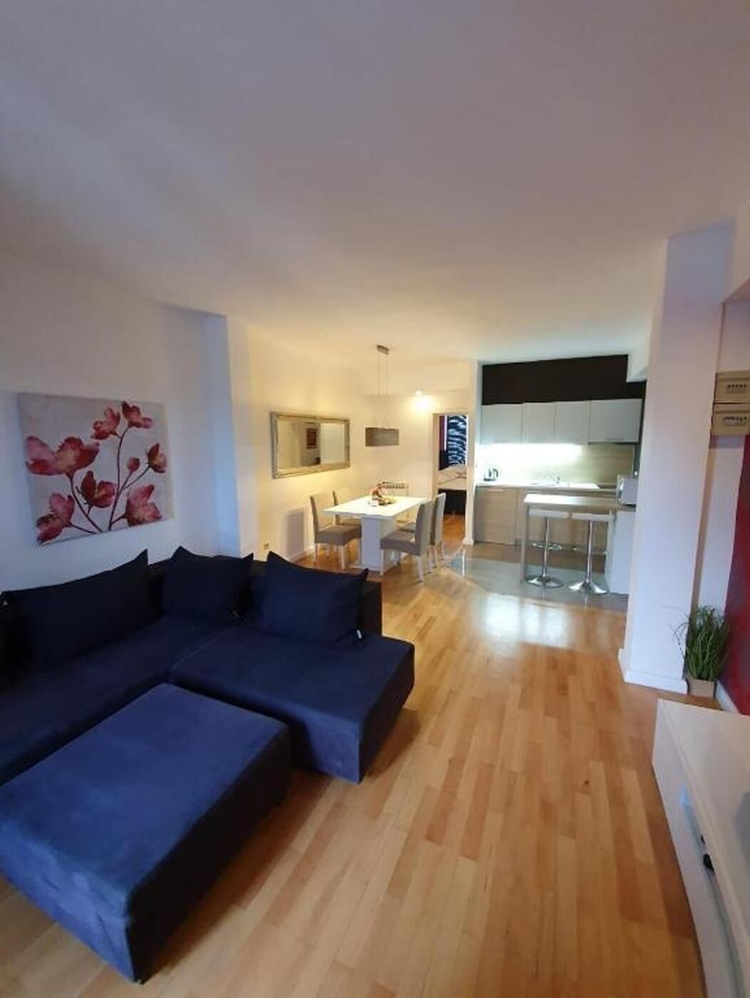 Una sala de estar moderna con un sofá seccional azul oscuro, un comedor con una mesa y sillas blancas, y una cocina al fondo.