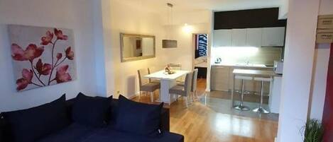 Una sala de estar moderna con un sofá seccional azul oscuro, un comedor con una mesa y sillas blancas, y una cocina al fondo.
