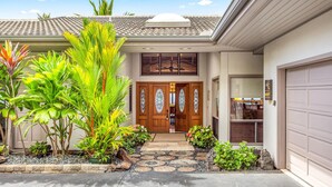 Una casa de dos pisos con porche delantero, garaje y un jardín bien cuidado con plantas tropicales.
