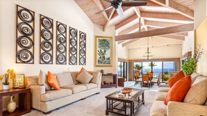 Un salón espacioso con techo alto, un ventanal con vistas al océano y un balcón con mobiliario de exterior.