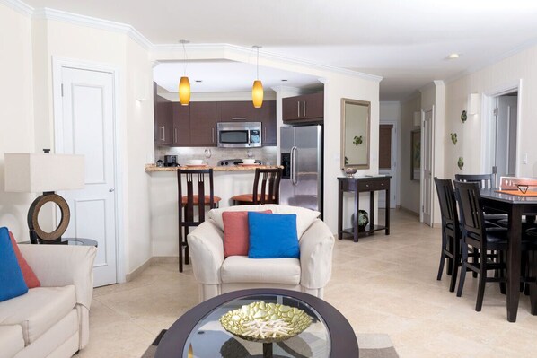 A modern kitchen with a microwave, refrigerator, and dining area. There is a white sofa with blue and red cushions, and a glass coffee table with decorative items.