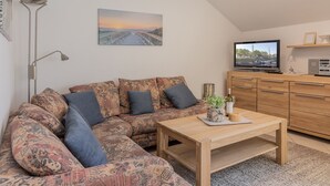 Een gezellige woonkamer met een hoekbank, een houten salontafel, een televisie en een schilderij van een ondergaande zon aan de muur.
