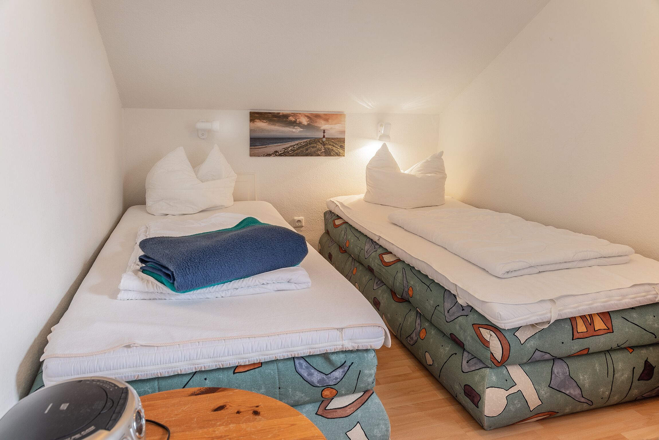 Una habitación pequeña y acogedora con dos camas individuales, cada una con una almohada y un edredón doblado. Hay una mesita con una silla, una lámpara de pared y un cuadro de un faro en la pared.