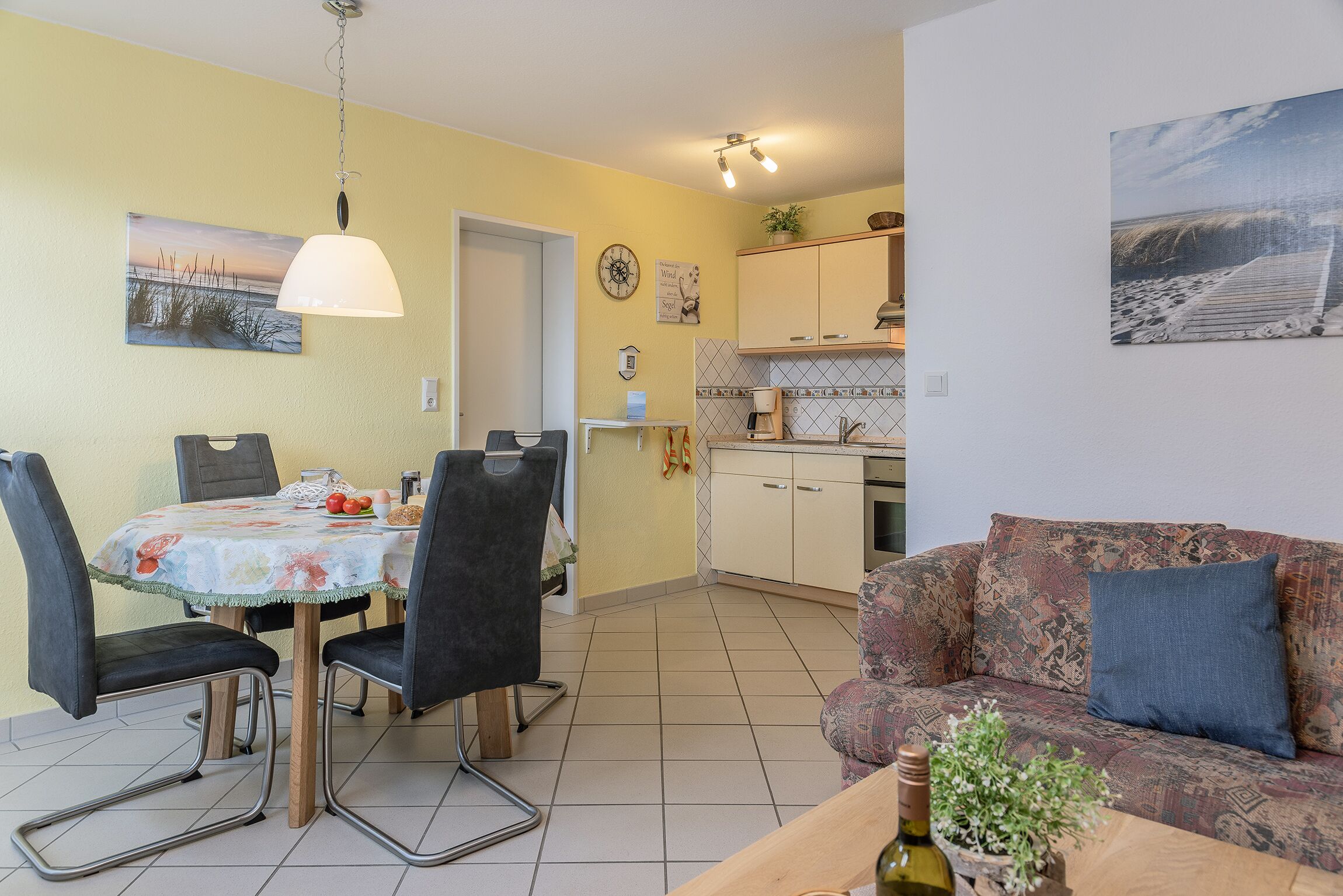 A dining area with a table and chairs, a kitchen in the background, and a sofa with a cushion.
