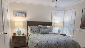 Une chambre à coucher avec un lit, deux tables de chevet, une lampe, un tableau au mur et une fenêtre avec des volets.