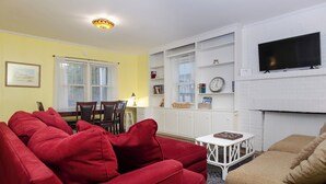 Ein Wohnzimmer mit einem roten Sofa, einem beigen Sofa und einem weißen Couchtisch. An der Wand ist ein Fernseher angebracht, ein Bücherregal steht dort, und ein Fenster mit Jalousien ist zu sehen.