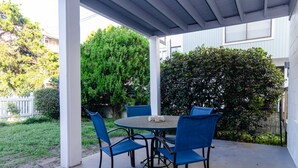 Eine überdachte Terrasse mit einem runden Tisch und blauen Stühlen, umgeben von Grünpflanzen und einem weißen Zaun.
