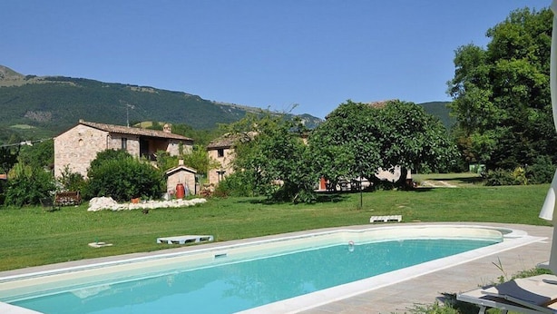 Una piscina con sillas de descanso, edificios de piedra y un área verde con árboles y colinas al fondo.