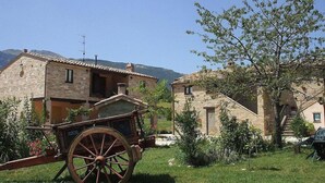 Un edificio de piedra con techo de tejas, un carro de madera y un árbol en un área cubierta de césped.