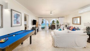 Ein Zimmer mit Pooltisch, einem weißen Sofa und Meerblick durch große Glasfenster.