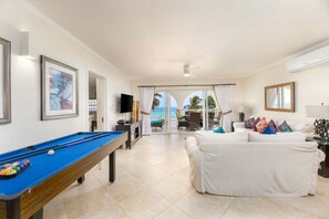 A room with a pool table, a white sofa, and a view of the ocean through large glass doors. - Luxury Oceanfront Condo with Pool - Sapphire 313 (Saint Lawrence Gap)