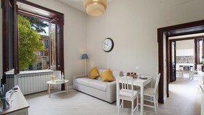 Un soggiorno moderno con un divano, un tavolino da caffè e un tavolo da pranzo. C'è un orologio al muro e una finestra con vista sugli edifici.