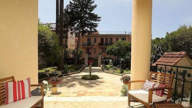 Un cortile con una panchina, una sedia e un tavolo di legno. Si vedono piante e un edificio sullo sfondo.