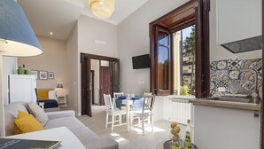 Un soggiorno moderno con divano, tavolo da pranzo con sedie, TV a parete e angolo cottura con rivestimento in piastrelle.
