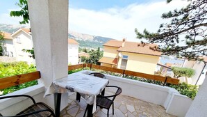 Un balcón con una mesa y sillas, suelo de piedra y vistas a casas y montañas.