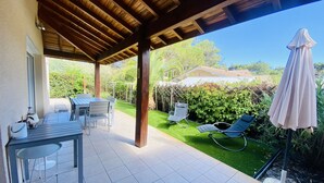 A covered patio area with a table and chairs, a lawn chair, and a covered umbrella. - Maison 4 chambres proche plage (023) (Biscarrosse)