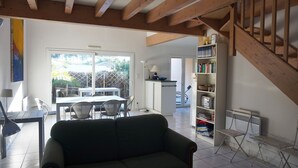 A living room with a sofa, a dining table with chairs, a bookshelf, and a staircase. - Maison 4 chambres proche plage (023) (Biscarrosse)