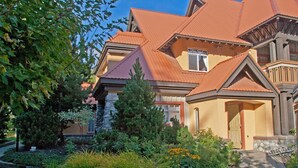 Une maison à deux étages, avec un toit rouge, des fondations en pierre et un jardin aménagé où poussent différentes plantes et fleurs.