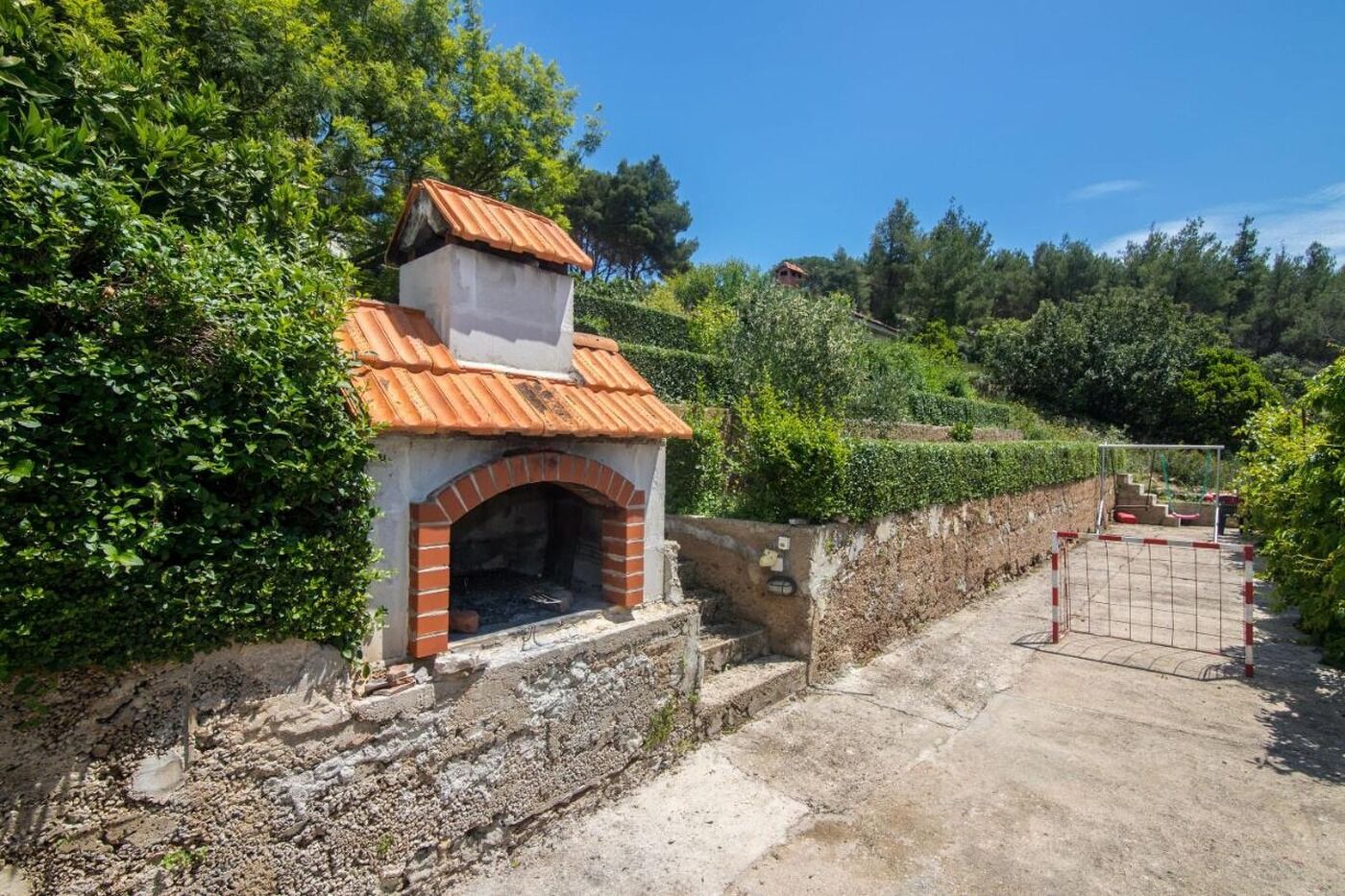 Ein Backofen aus Ziegelsteinen im Freien mit einem gefliesen Dach, umgeben von üppiger Bepflanzung und einer Steinmauer.