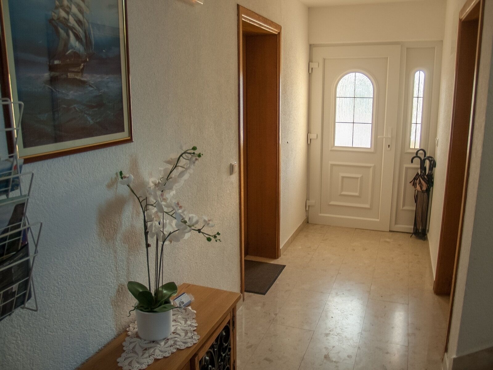 A hallway with a potted orchid on a wooden console table, a framed picture on the wall, and a door with a window.