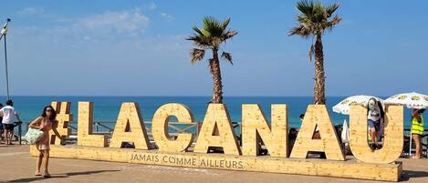 A wooden sign with the text "LAGANAU" and "JAMAIS COMME AILLEURS" on a beach with palm trees and the sea in the background.