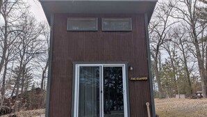 A small, single-story building with a dark wooden exterior, a large sliding glass door, and a sign that reads 'THE GLAMPER'.