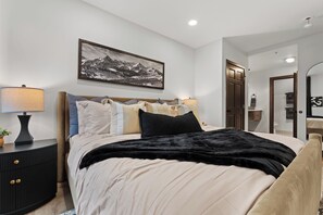 Une chambre à coucher comprenant un lit, des tables de chevet, une commode, une lampe fixée au mur, un tableau encadré et une porte donnant sur une autre pièce.