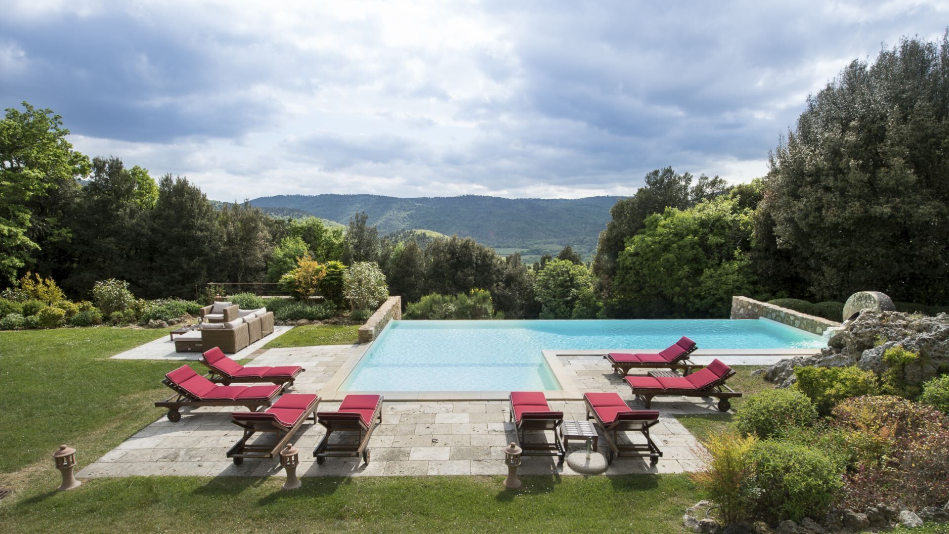 A rectangular swimming pool with a clear blue water surface, surrounded by a stone deck. There are multiple lounge chairs with red cushions arranged around the pool. The pool area is set against a backdrop of lush greenery and distant hills.