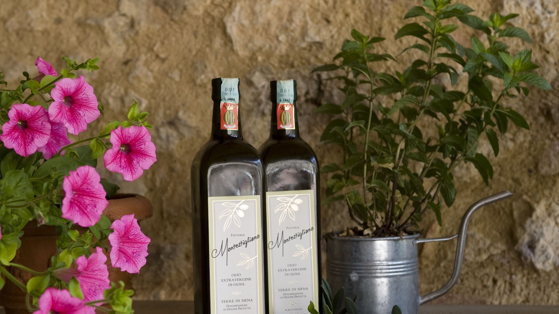 Two bottles of Montestigliano extra virgin olive oil with a metal watering can and pink flowers in the foreground.