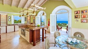 Un'ampia cucina con una grande isola, mobili in legno e vista sull'oceano. È presente una zona pranzo con un tavolo in vetro e sedie in vimini.
