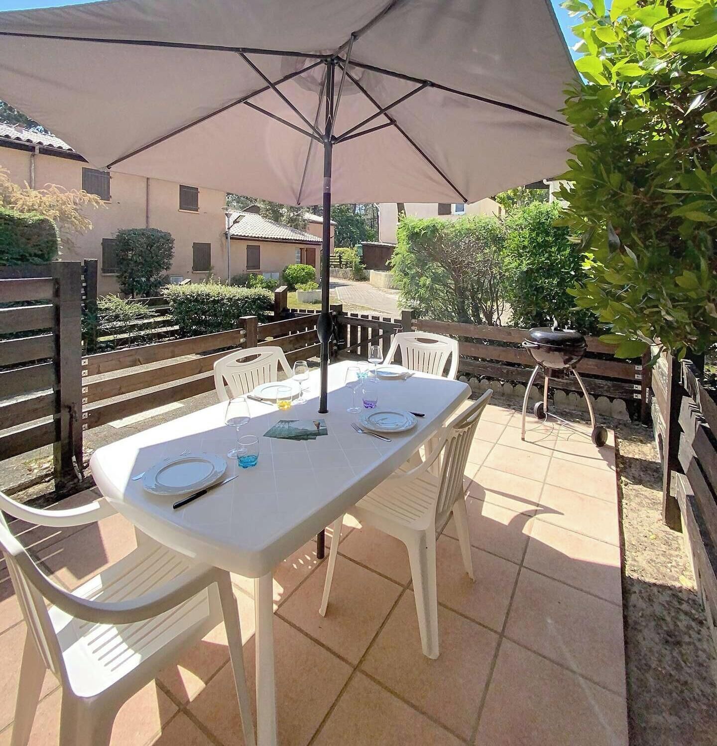 Eine Terrasse im Freien mit einem für vier Personen gedeckten Tisch, umgeben von weißen Plastikstühlen, unter einem großen Sonnenschirm.