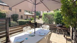 Un patio al aire libre con una mesa preparada para cuatro personas, rodeada de sillas de plástico blancas, bajo un paraguas grande.