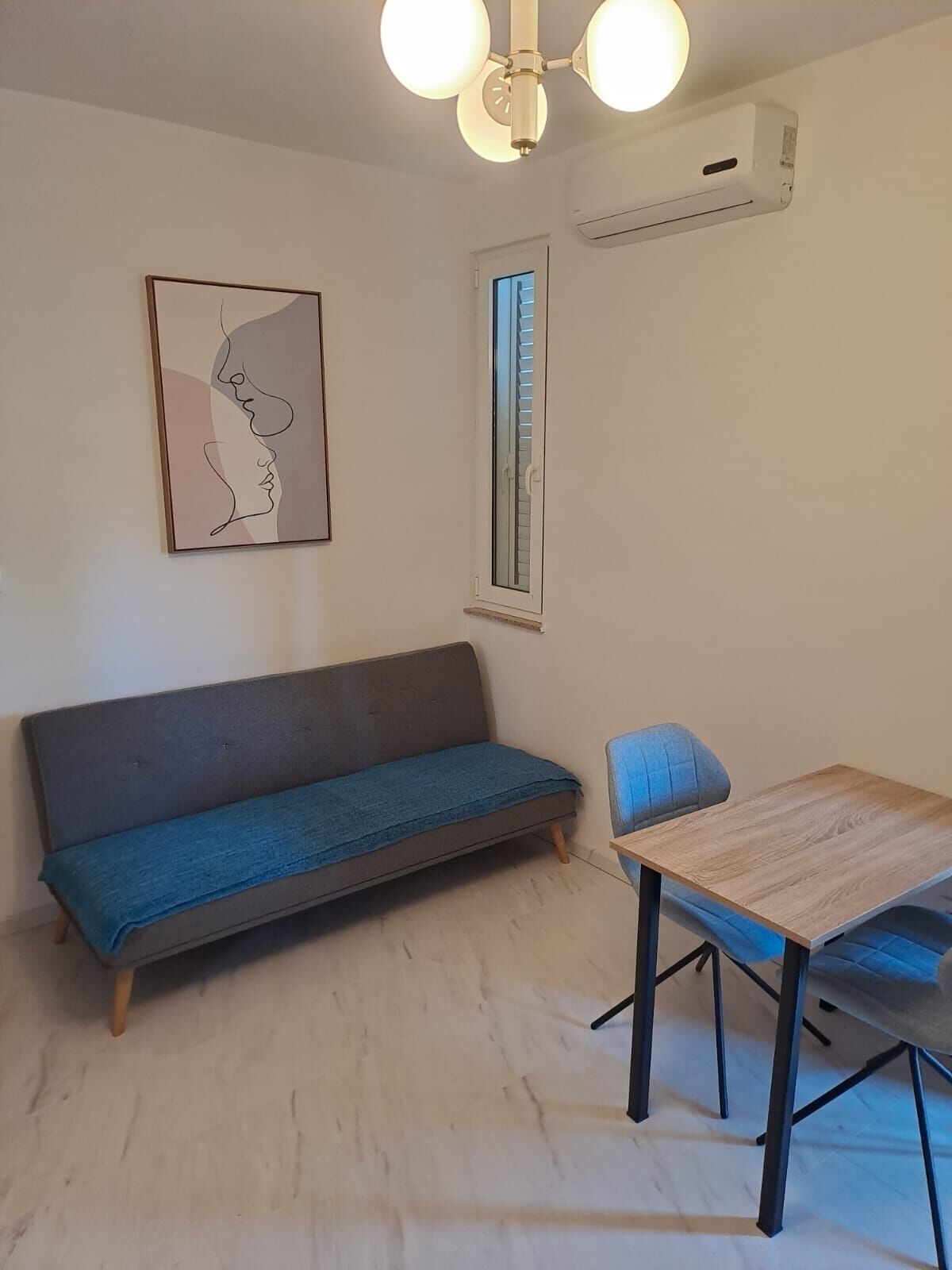 A room with a sofa, a table, a chair, a ceiling fan, an air conditioning unit, and a window with blinds.