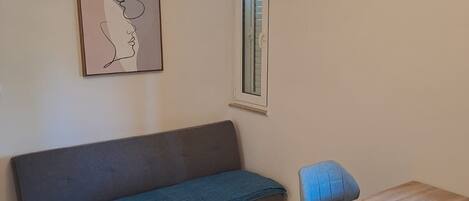A room with a sofa, a table, a chair, a ceiling fan, an air conditioning unit, and a window with blinds.