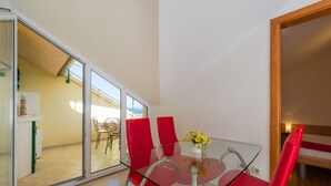 Un comedor con mesa de vidrio y sillas rojas, un florero con flores amarillas, y vistas a una zona de estar exterior a través de un ventanal.