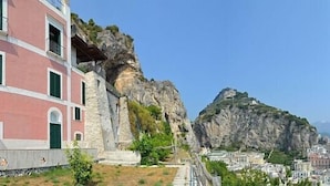 Un edificio con facciata rosa, persiane verdi e base in pietra. Si trova su un pendio con una scogliera rocciosa sullo sfondo.