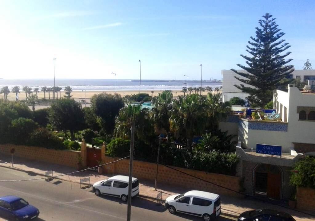 Un hôtel avec une enseigne indiquant « Hôtel Villa Quiera », situé près d’une plage bordée de palmiers, sous un ciel dégagé.