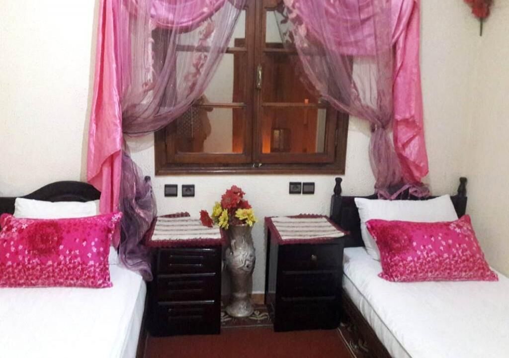 Une chambre avec deux lits, une fenêtre avec des rideaux, un vase de fleurs et une petite table placée entre les lits.
