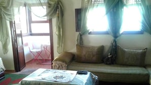 Ein Wohnzimmer mit einer Couch, einem kleinen Tisch und einem Sessel. Ein Fenster mit Vorhängen und Ausblick.