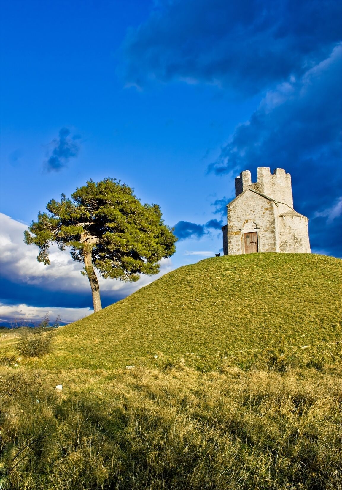 Un bâtiment en pierre, surmonté d’une tour, se dresse sur une colline verdoyante, sous un ciel dégagé.