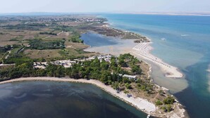 Vista aérea de uma área costeira com uma praia, um estacionamento e um pequeno conjunto de edifícios rodeados por árvores.