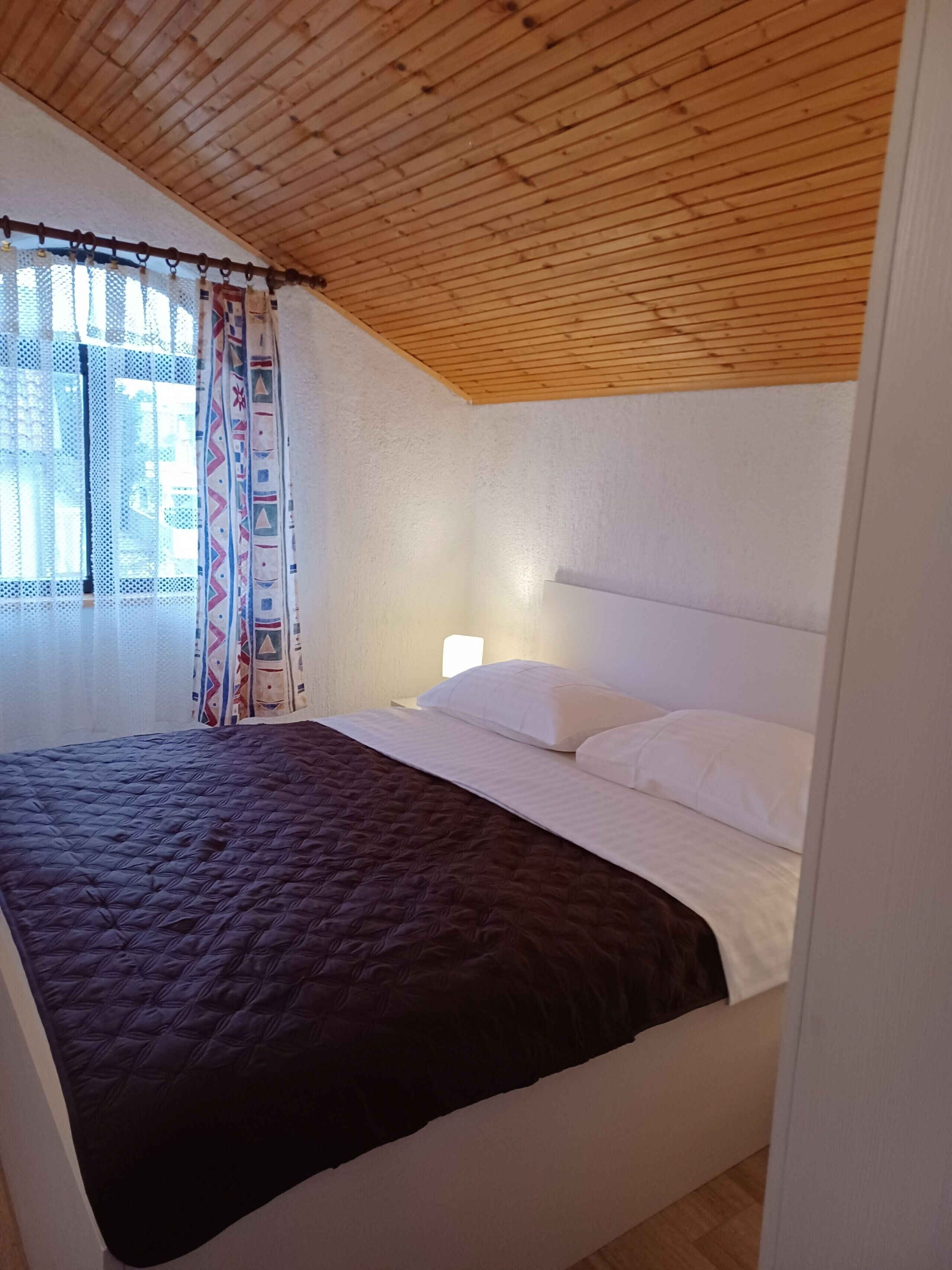 Une chambre avec un plafond en bois, un lit avec une literie blanche et marron, une table de chevet et une fenêtre ornée de rideaux à motifs.