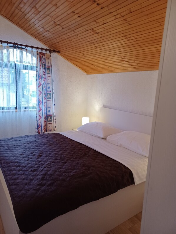 Une chambre avec un plafond en bois, un lit avec une literie blanche et marron, une table de chevet et une fenêtre ornée de rideaux à motifs.