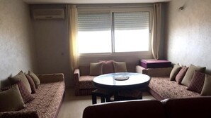 Een woonkamer met een zithoek, een rond salontafeltje en een raam met jaloezieën.
