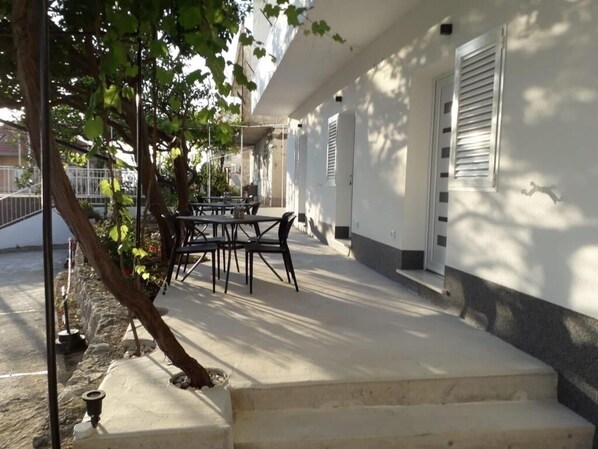 A covered outdoor seating area with a table and chairs, a tree, and a staircase leading to a building with white walls and shutters.