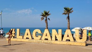A wooden sign with the text "LAGANAU" and "JAMAIS COMME AILLEURS" on a beach with palm trees and the sea in the background.