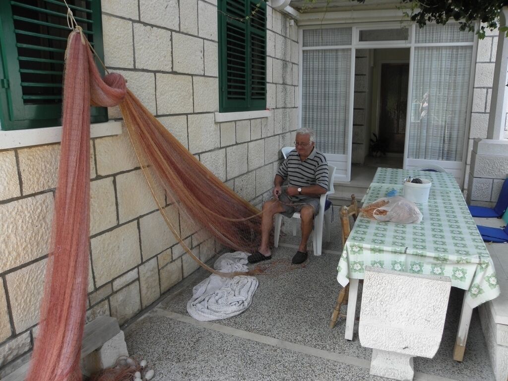 Un hombre sentado en una silla en un área exterior techada, con una hamaca, una mesa con mantel y una ventana con contraventanas verdes.