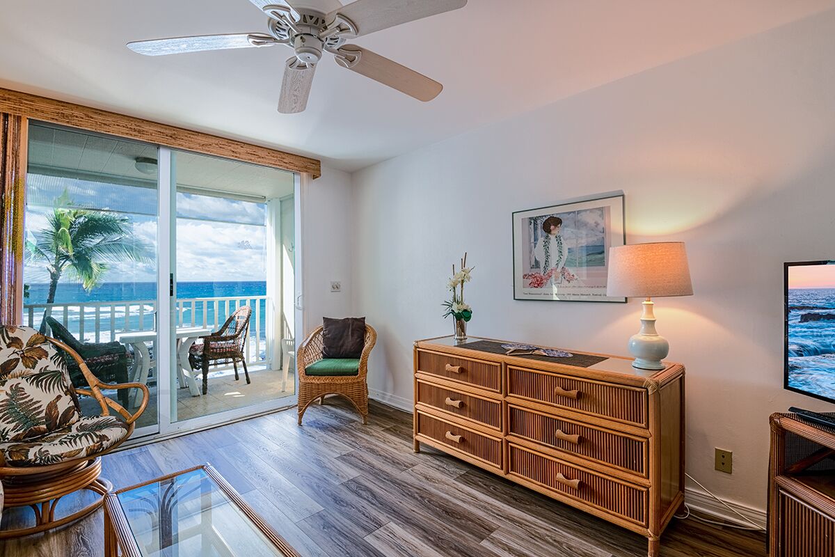 A living room with a view of the ocean, a ceiling fan, wicker furniture, a wooden cabinet, and a framed picture on the wall.