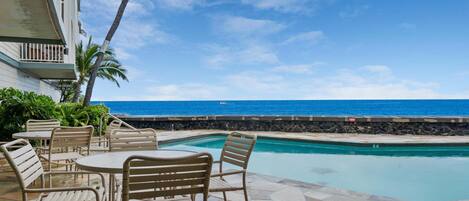 A poolside area with chairs and tables, a stone wall, and a view of the ocean.